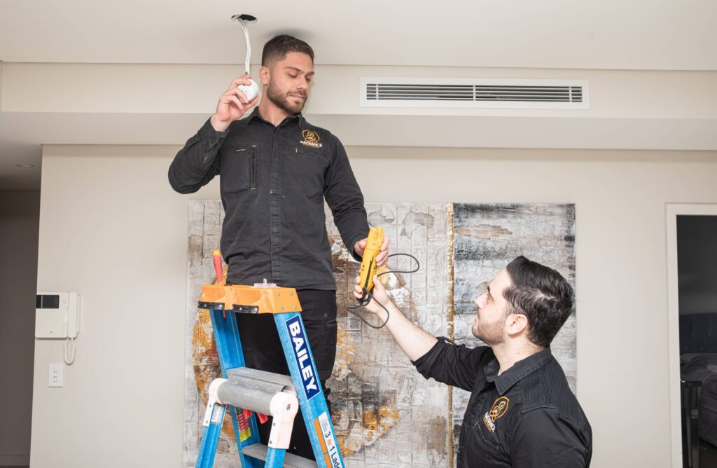 Two men installing a ceiling fan in a room, carefully aligning the blades and securing the wiring.