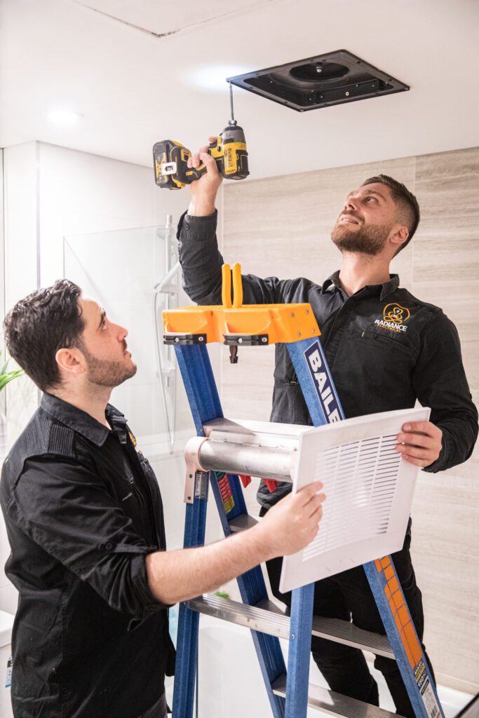 Two men wearing protective gear are working on a bathroom ceiling, using tools and materials to repair or renovate the area.